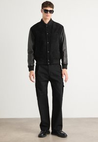 Black suede bomber jacket with leather sleeves, ribbed collar and cuffs, featuring dark buttons. Paired with black cargo pants and shoes.