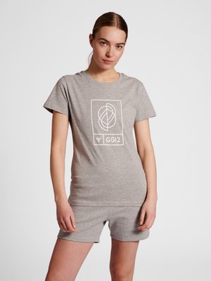 Kvinde med brunt hår bundet tilbage iført lysegrå T-shirt og matchende shorts, stående mod en ensfarvet hvid baggrund.