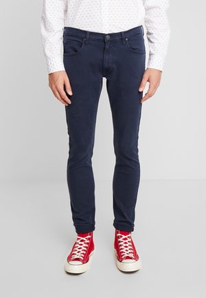 Mann trägt dunkelblaue schmale Jeans, weißes gemustertes Hemd und rote High-Top-Sneaker und steht auf einem hellgrauen Boden.
