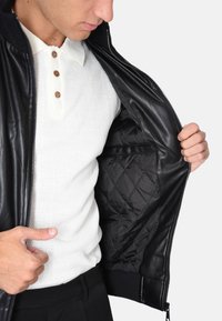 Giacca in pelle nera con zipper, fodera interna trapuntata e polsi a coste. Il modello indossa un polo bianco a trama sotto.