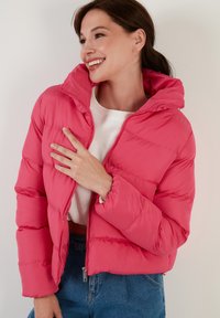 Ružová pufová bunda s kapucňou, s prešívaným dizajnom a predným zipsom. Nosí sa cez biely top a modré džínsy.