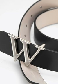 Zwarte leren riem met een grote metalen zilveren "W" gesp. De binnenkant is glad beige met stiksels en meerdere gaatjes.