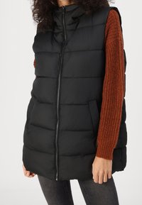 Veste sans manches matelassée noire avec un col haut et une fermeture éclair, dotée d'une manche tricotée de couleur rouille contrastante sur le côté droit.