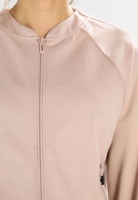 Giacca zip-up rosa chiaro con scollatura arrotondata, maniche raglan e tasche laterali con dettagli in hardware nero. Tessuto morbido e testurizzato.