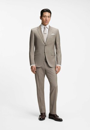 Man staat, draagt een beige pak met een wit overhemd, een patroon stropdas, een pochet en donkerbruine leren nette schoenen op een effen achtergrond.