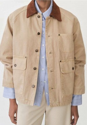 Veste beige avec col en velours côtelé marron, boutons-pression métalliques et trois poches avant portée sur une chemise bleu clair à boutons et un pantalon beige.