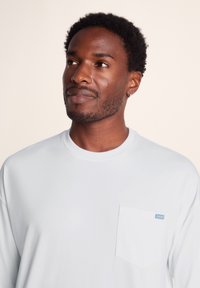 Long-sleeve white T-shirt made of soft fabric, featuring a chest pocket with a small blue tag. Smooth texture and relaxed fit.