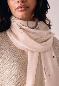 Sciarpa beige con motivi in filo dorato, realizzata in un tessuto leggero. Indossata sopra un maglione lavorato a maglia beige testurizzato. Morbida caduta e aspetto leggero.