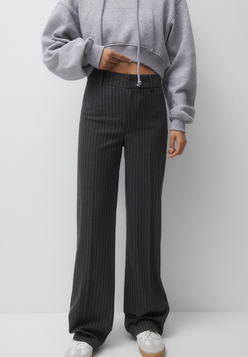Sudadera gris corta con un bolsillo frontal, combinada con pantalones de tiro alto negros a rayas. Footwear presenta zapatillas ligeras gris claro con detalles blancos.