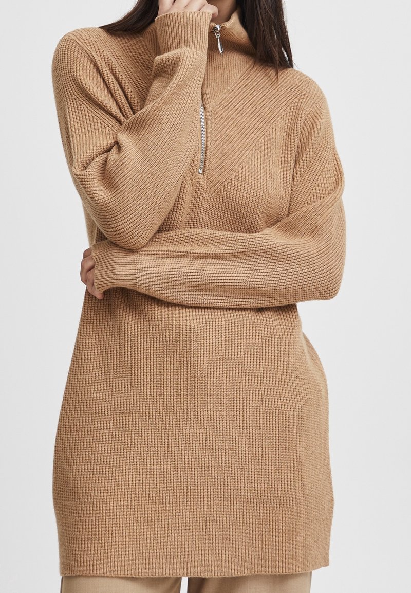 Maglione lavorato a maglia in marrone chiaro con colletto alto, chiusura con zip e texture a costine; presenta dettagli a pannello diagonale sul petto.