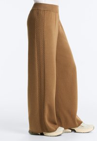 Rjave široke pletene hlače z navpičnim teksturiranim vzorcem na strani, nošene z belimi supergami.