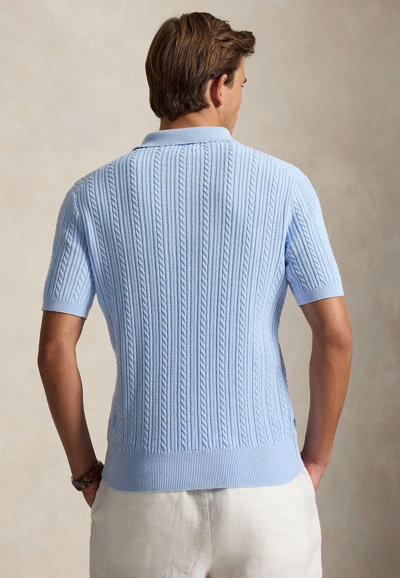 Homme portant un polo bleu clair à manches courtes en maille torsadée et un pantalon blanc, debout, mains dans les poches, vu de dos.