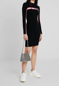 Vestito lungo a maniche lunghe nero a coste con strisce rosse e bianche, caratterizzato da un logo sul petto. Abbinato a una borsa in rete metallica e sneakers bianche.