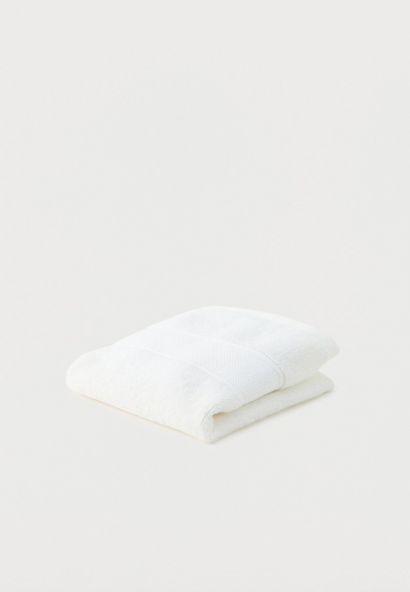 Opgevouwen witte badhanddoek met gestructureerde band, geplaatst op een effen wit oppervlak.