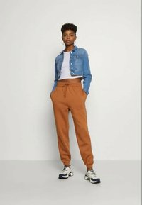 Casaco de ganga na cor azul clara, com um design cropped, combinado com calças de fato de treino cor caramelo com cintura elástica e bolsos, usadas com ténis brancos e pretos.