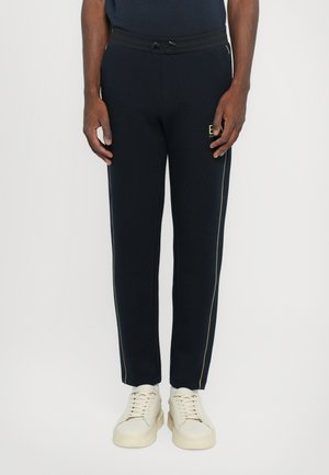 Pantalons de survêtement navy avec une coupe fuselée, taille élastique, cordon de serrage et rayures dorées sur les côtés. Portés avec des baskets de couleur claire.