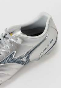 Cizma de fotbal argintie cu un suprafață texturată, având accente albastre, un design elegant și cu numele "MONARCIDA NEO" pe lateral.