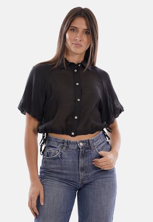 Donna con capelli castani lisci indossa un top corto nero con bottoni e jeans blu a vita alta su uno sfondo semplice.