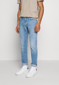 Světle modré džíny s rovnýma nohavicema, mírně ohrnuté manžety, spárované s bílými teniskami a béžovým tričkem.