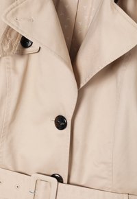 Trench beige avec larges revers, boutons noirs, patte d'épaule boutonnée et ceinture assortie à la taille.