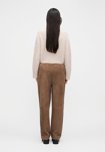 Bézs kötött pulóver és barna szarvasbőr nadrág. A pulóver hosszú ujjú; a nadrág bőszárú és sima tapintású. Fekete cipő.