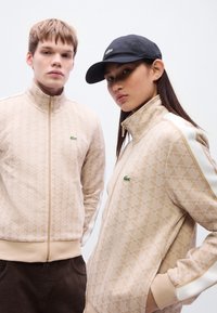 Μπεζ μπουφάν με φερμουάρ και γεωμετρικά σχέδια, με λευκές πλευρικές ρίγες και μικρό πράσινο λογότυπο. Μαύρο καπέλο φορεμένο από το μοντέλο στα δεξιά.