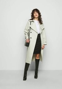 Sieviete ar pūkainiem matiem, ģērbusies gaismā trenčkotā virsējā apģērbā pār baltu topu un melnām svārkiem, tur melnu plecu somu un valkā melnas augstus zābakus.