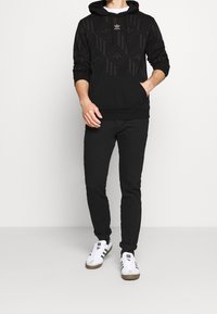 Černá mikina s kapucí s geometrickým vzorem a klokáním kapsou, spárovaná s černými úzkými kalhotami a bílými teniskami s černými pruhy.