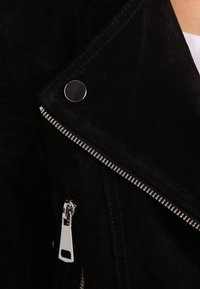 Černá semišová motorkářská bunda se stříbrnými prvky, která má výrazný zip, detaily na patentkách a texturovaný povrch.