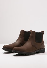 Bottes Chelsea en cuir marron avec des panneaux latéraux élastiques, un bout arrondi et une semelle extérieure en caoutchouc texturée pour une meilleure adhérence. Finition lisse avec des détails minimaux.