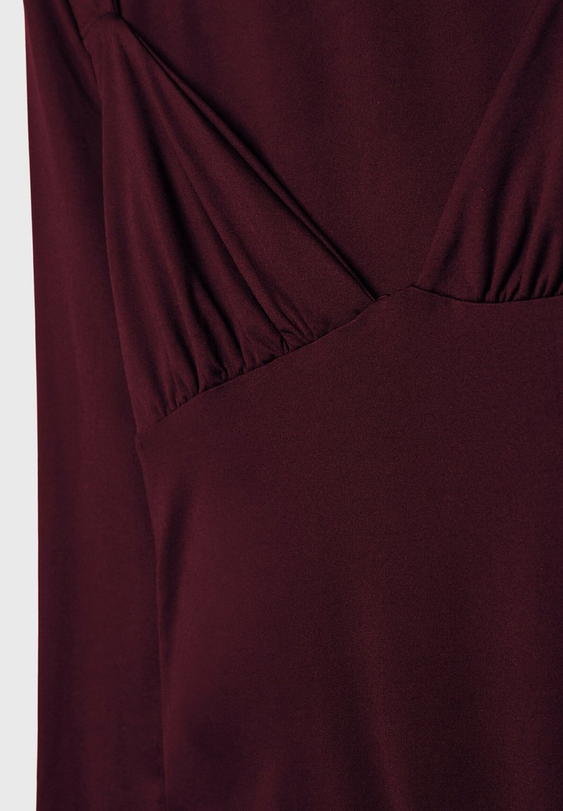 Tessuto di abito bordeaux con dettagli sulle cuciture; trama morbida e elastica; presenta delle pieghe lungo il busto per dare forma e una silhouette leggermente aderente.