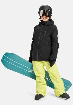 Enfant portant un casque noir, des lunettes, une veste noire, un pantalon de neige jaune et des gants, tenant un snowboard turquoise.