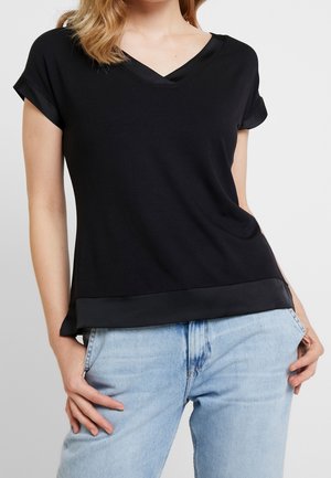 Femme portant un haut noir à manches courtes en V avec une bordure en satin, associé à un jean en denim bleu clair, debout avec la main dans la poche.