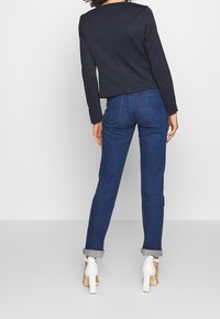 Marineblauwe blazer met lange mouwen, gecombineerd met donkerblauwe jeans. De jeans hebben een opgerolde zoom; witte hoge hakken maken de outfit af.