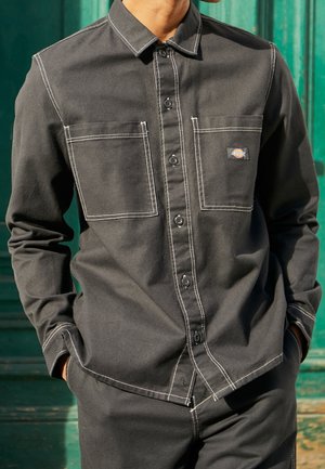 Man wearing dark gray button-up shirt with white stitching and two front chest pockets, standing with hands in pockets against green background.