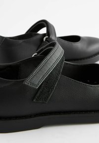 Chaussures en cuir noir avec un bout arrondi, des sangles élastiques et un détail texturé sur les sangles. Semelle plate et accents de couture minimaux.
