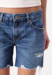 Modré džínové kraťasy s třepeným lemem, opotřebovanými detaily a oranžovým šitím. Mají standardní rozložení předních knoflíků a kapes.
