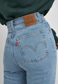 Jean en denim bleu clair à taille haute, avec un patch en cuir marron, une étiquette en ruban rouge et un motif de couture distinctif sur la poche arrière.
