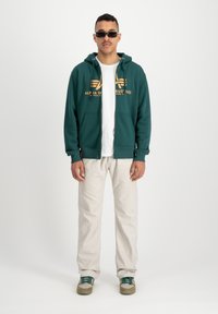 Sudadera verde con cremallera y logo naranja, llevada sobre una camiseta blanca, combinada con pantalones de colores claros y zapatos con detalles verdes.