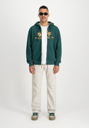 Felpa verde con zip e logo arancione, indossata sopra una maglietta bianca, abbinata a pantaloni chiari e scarpe con dettagli verdi.