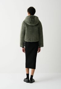 Groene met bont gevoerde cropped jas met capuchon, gecombineerd met een zwarte geribbelde midi rok en zwarte schoenen. De stof lijkt zacht en textuur te hebben.