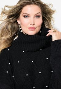 Schwarzer Strickpullover mit hohem Kragen, der ein Rautenmuster und Perlenakzente aufweist. Model zeigt Ohrringe und einen Ring.
