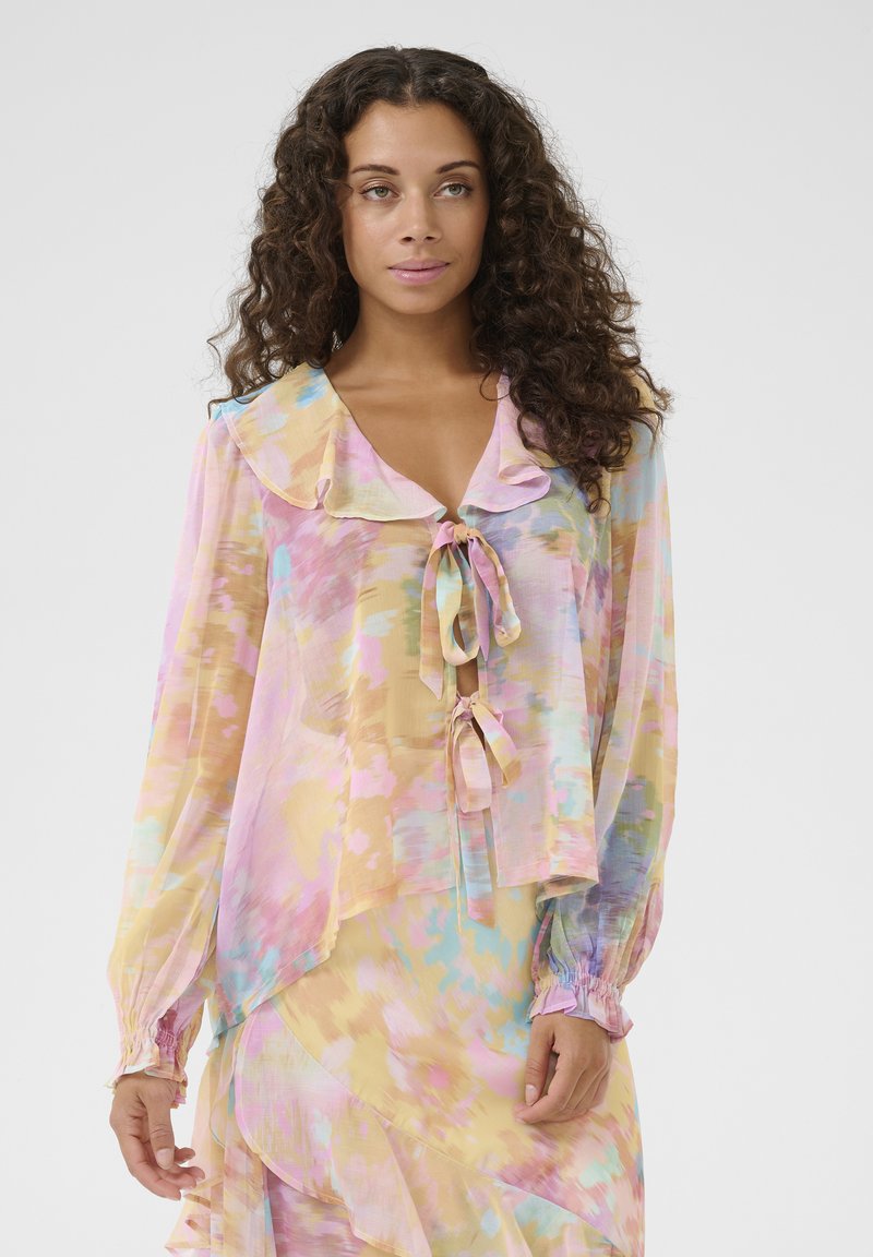 Blusa trasparente e multicolore con morbide tonalità pastello, colletto arricciato, chiusura con fiocco sul davanti e polsini elasticizzati. Leggera con un design fluido.