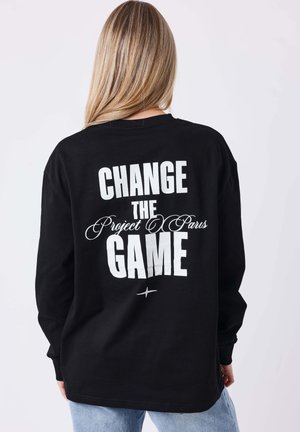 Femme aux longs cheveux blonds portant un t-shirt noir à manches longues avec un texte blanc "CHANGE THE Project Paris GAME" dans le dos, associé à un jean bleu clair.