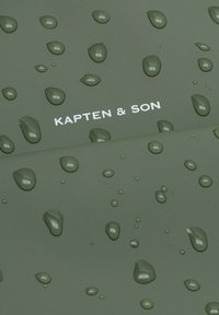Waterafstotende stof in donkergroen met waterdruppels; het logo "KAPTEN & SON" in het wit prominent weergegeven.