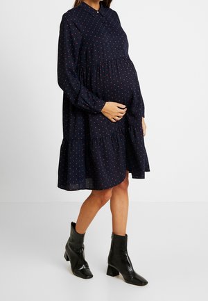 Gravid kvinde iført en mørkeblå kjole med røde polka prikker og sorte ankelstøvler står mod en ensfarvet baggrund.