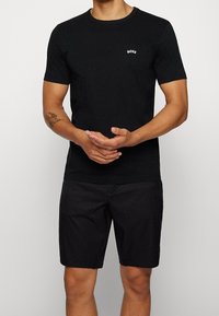 Homme portant un T-shirt noir à manches courtes avec le logo "BOSS" sur la poitrine et un short noir, debout les mains jointes devant lui.
