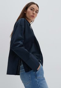 Camicetta blu navy con bottoni, maniche lunghe, fessure laterali e taglio dritto. Indossata con jeans in denim azzurri. Tessuto liscio e leggero.