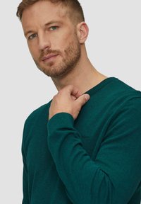 Türkis gestrickter Pullover mit Rundhalsausschnitt, gerippten Bündchen und einem dezenten Logo-Detail. Weicher Stoff und lässige Passform.