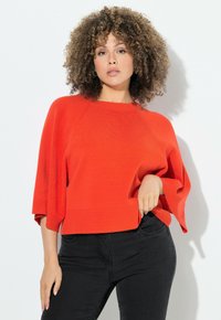 Roter, übergroßer Strickpullover mit geripptem Saum und halblangen �Ärmeln, kombiniert mit schwarzen Slim-Fit-Jeans. Glatte Textur und bequeme Passform.